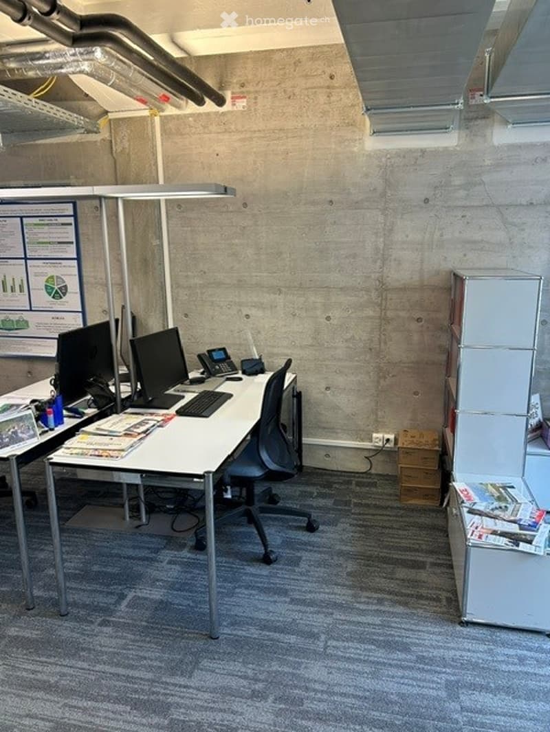 Modern office space with a white desk, black office chair, computer monitors, keyboard, telephone, and filing cabinets. Exposed concrete walls and industrial-style lighting.