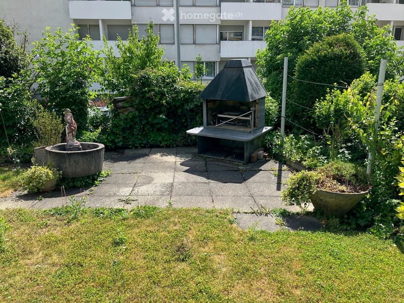 Garden area with a small barbecue, a fountain, and lush greenery, surrounded by a paved patio and a multi-story building in the background.
