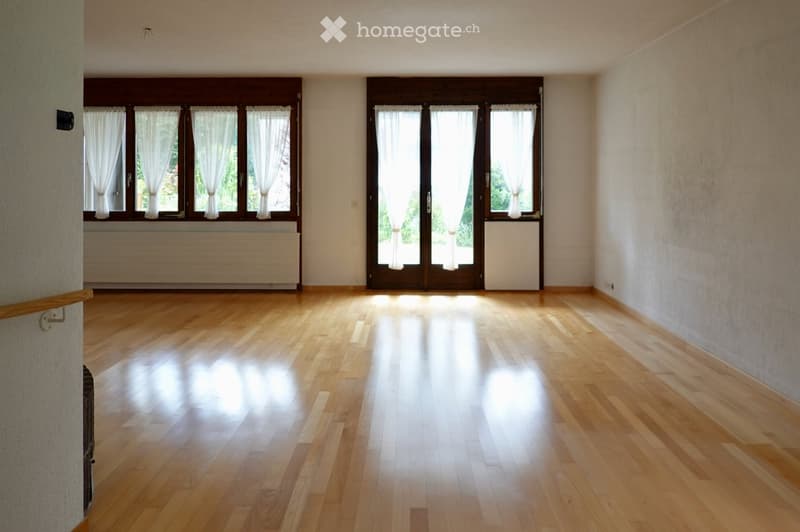 Empty room with wooden flooring, large windows with white curtains, and a radiator under the windows. The space is well-lit with natural light.
