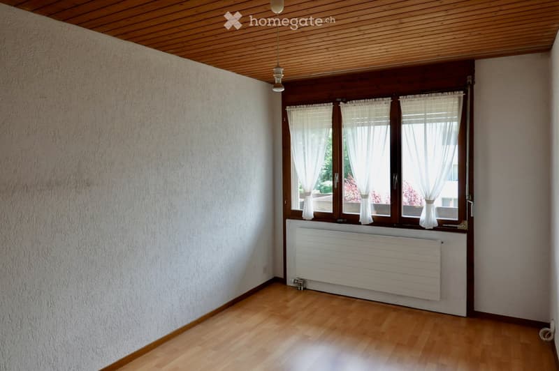 Empty room with wooden flooring, white walls, large windows with white curtains, and a white radiator below the windows.