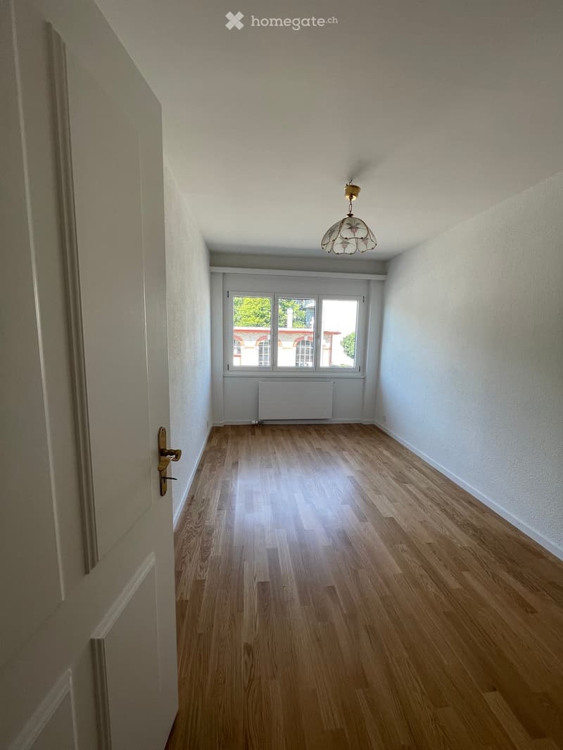 Empty hallway with wooden flooring, white walls, a single window at the end, and a decorative ceiling light fixture.