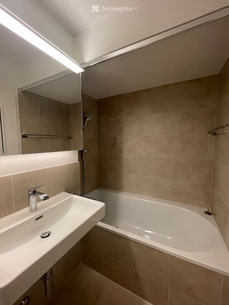 Modern bathroom with a white sink, chrome faucet, bathtub, and large mirror above the sink. Beige tiles cover the walls and floor.