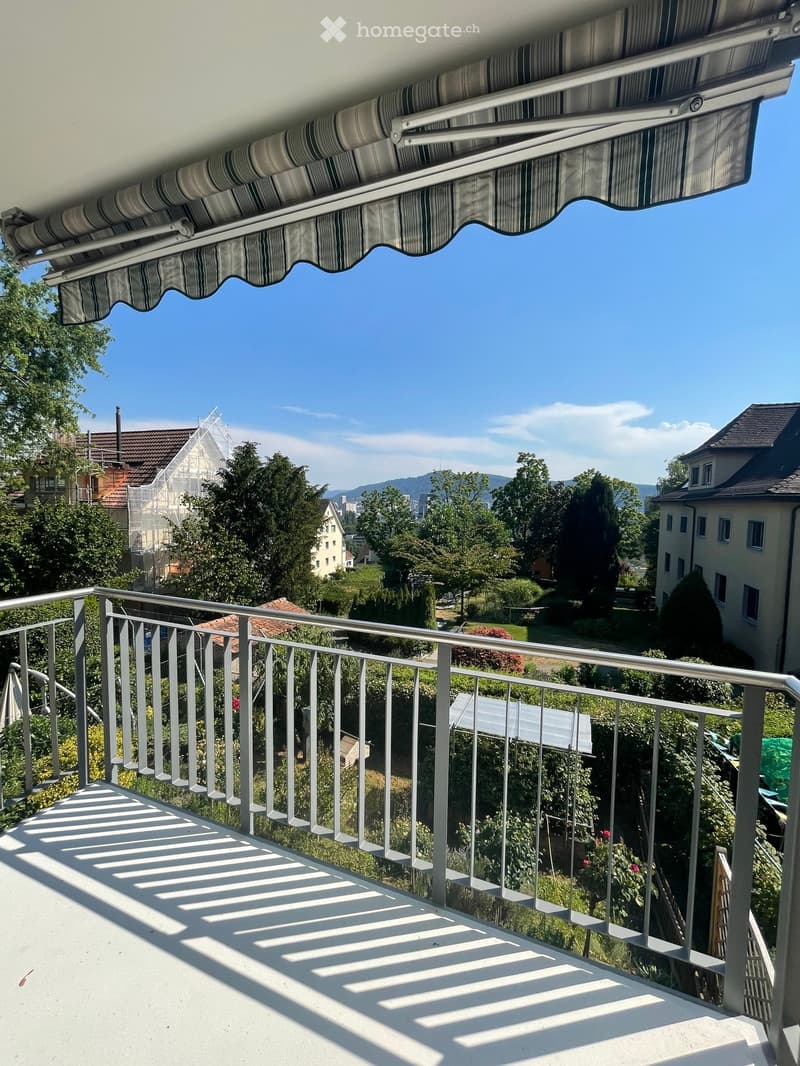 Balcony with metal railing, retractable awning, and scenic view of trees and houses.