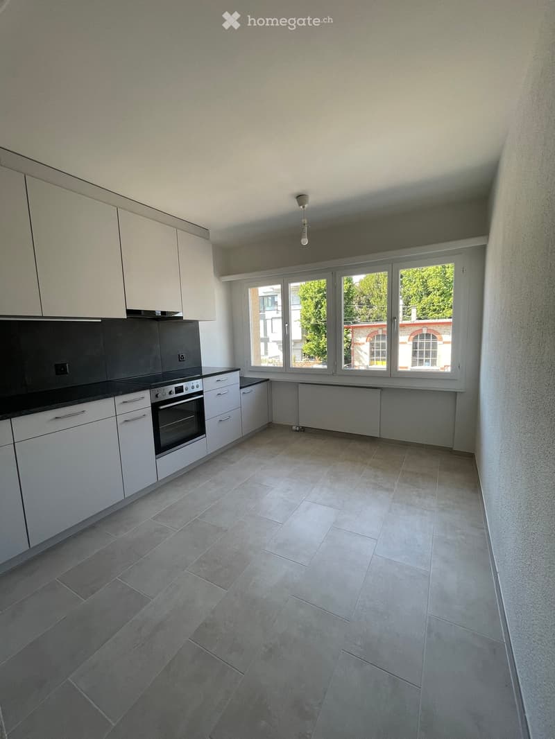 Empty kitchen with white cabinets, stainless steel oven, tiled floor, and large windows overlooking a green area.
