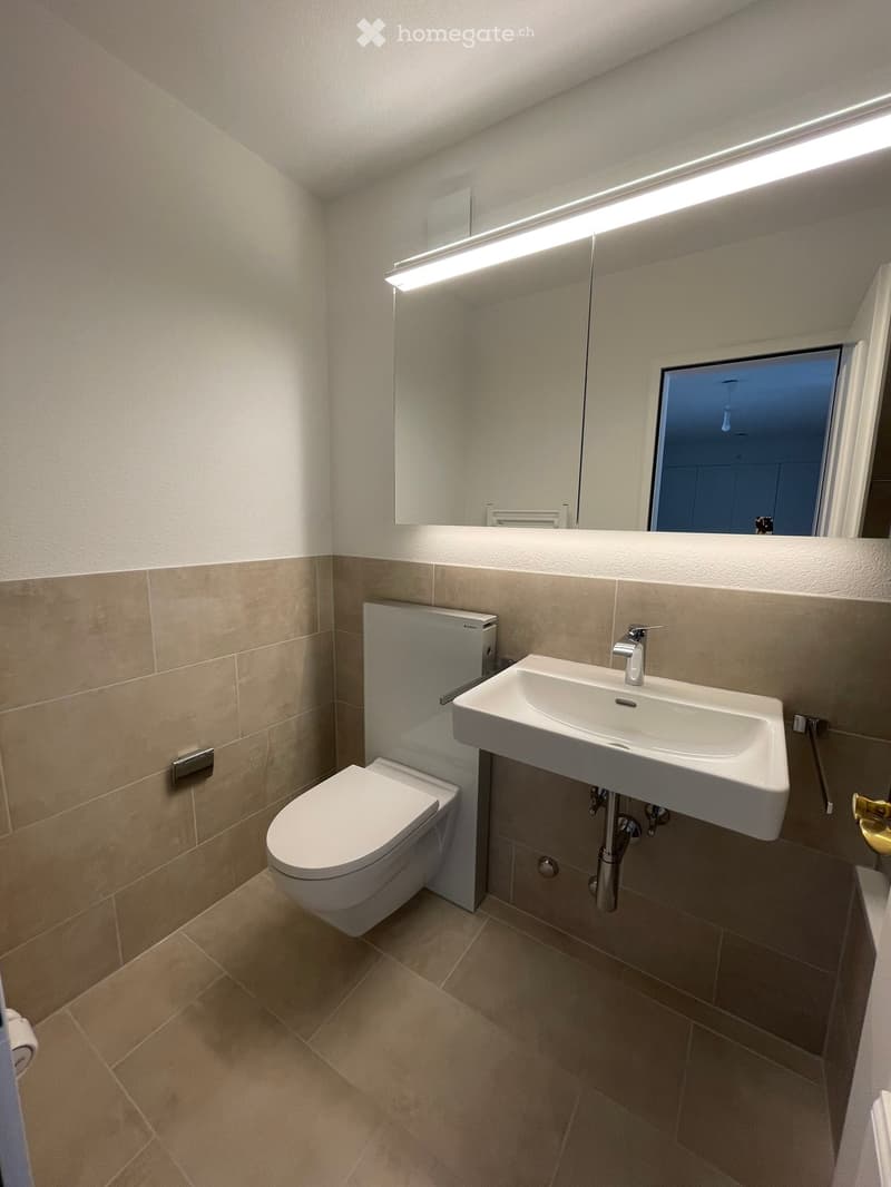 Modern bathroom with beige tiles, a white toilet, a white sink with a chrome faucet, and a large mirror with built-in lighting.