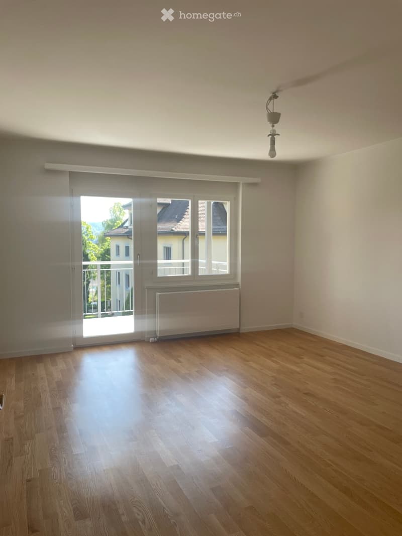 Empty room with wooden flooring, large window with blinds, radiator, and a hanging light fixture.