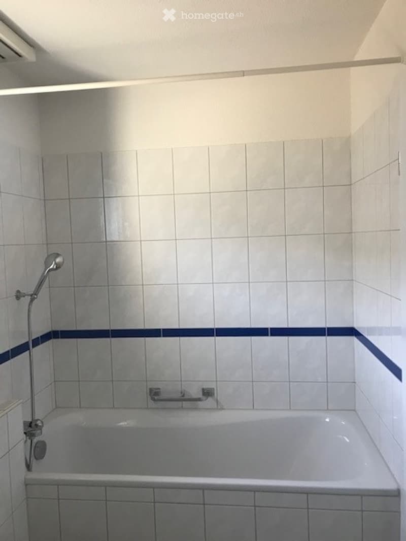 Clean, modern bathroom with white tiled walls and floor, a bathtub, a showerhead, and a towel rack.