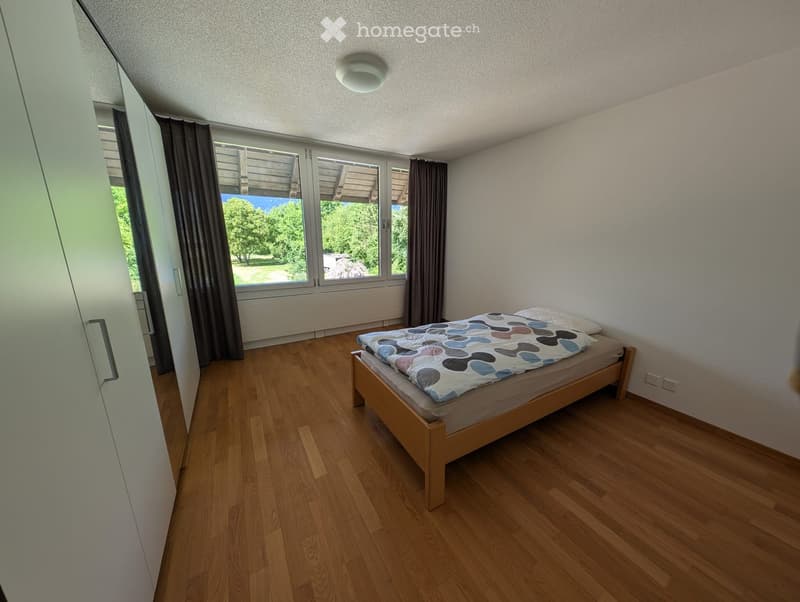 Minimalist bedroom with wooden flooring, a single bed with a colorful quilt, and a large window with curtains offering a view of greenery.
