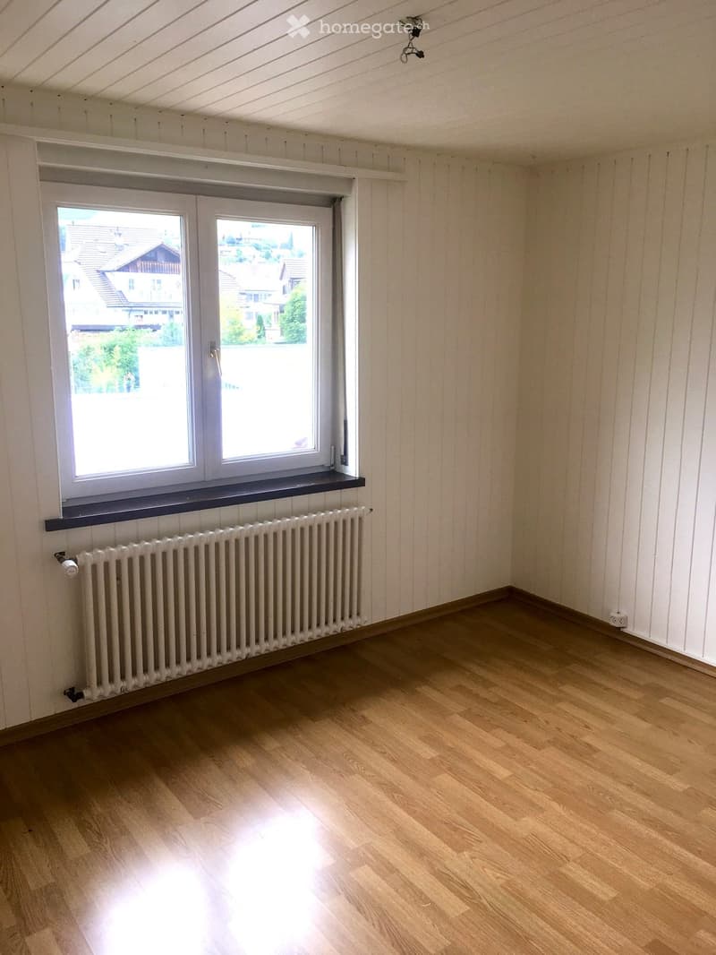 Empty room with wooden flooring, white paneled walls, a radiator, and a large window with a view of neighboring houses.