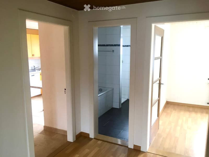Empty hallway with wooden flooring, leading to a bathroom and a kitchen area. The bathroom has white tiles and a bathtub, while the kitchen features light-colored cabinets and a sink.
