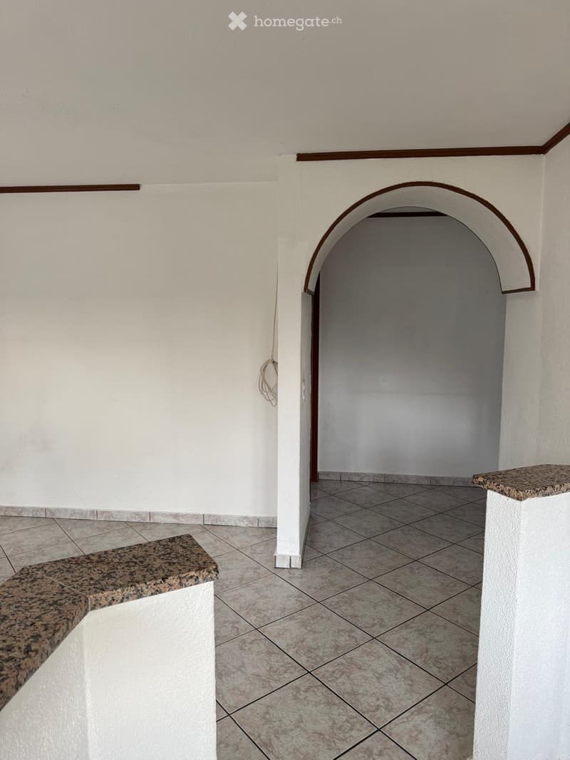Empty hallway with tiled floor, white walls, and an arched doorway leading to another room. Granite benches on either side.