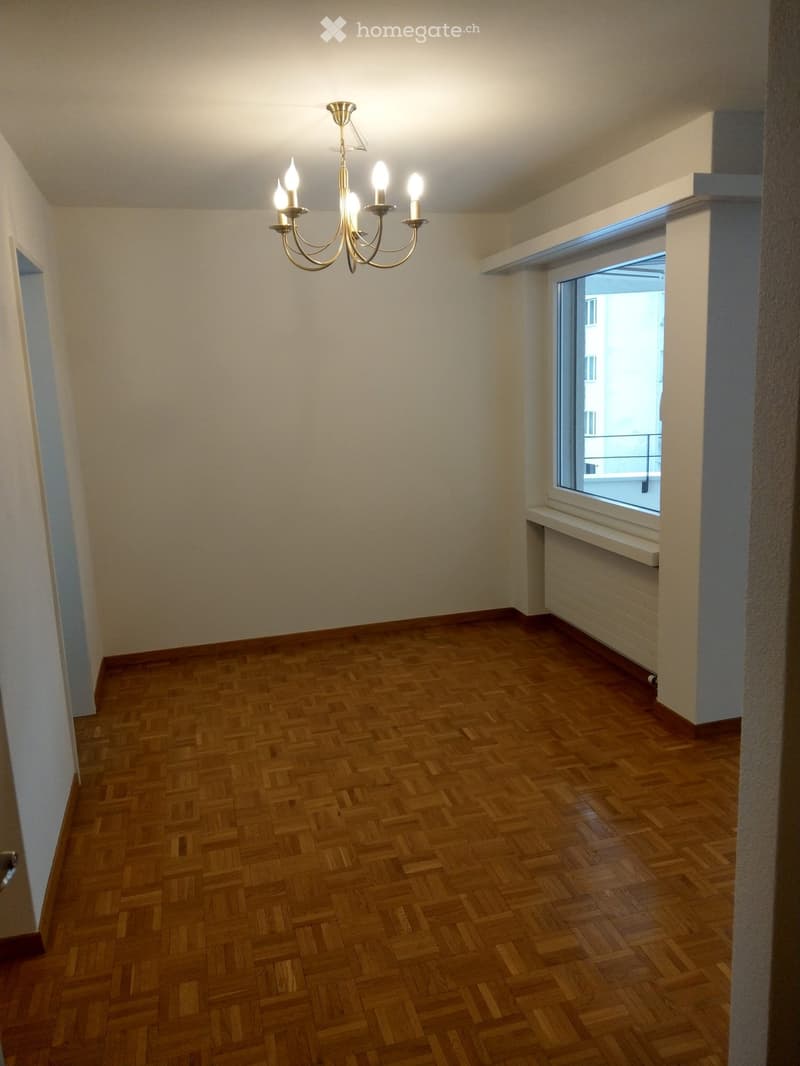 Empty room with wooden parquet flooring, white walls, a chandelier with multiple lights, and a window with a radiator below it.
