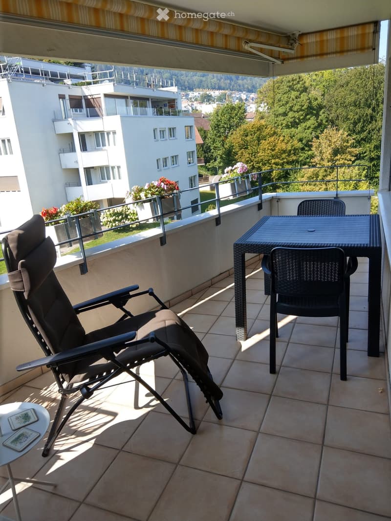 Balcony with tiled floor, table, chairs, and a view of the city and greenery.