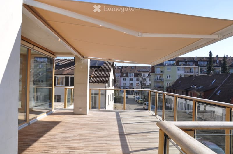 Balcony with wooden decking, glass railing, and retractable awning, overlooking a residential street with houses and trees.
