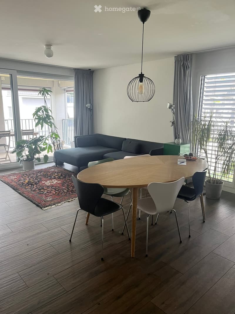 Modern living and dining area with a dark grey sofa, round wooden dining table, chairs, pendant light, side table, and plants near large windows and balcony access.