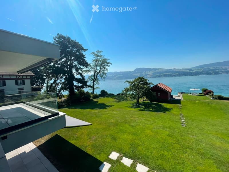 Lush green lawn with a modern glass balcony, a small red shed, and a scenic view of a lake and mountains in the background.