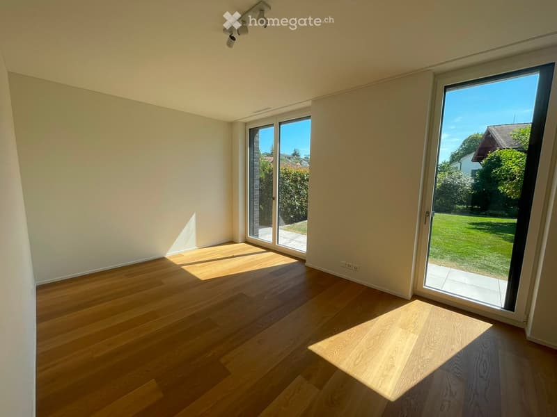 Empty room with wooden flooring, large sliding glass doors leading to a garden, and natural light streaming in.