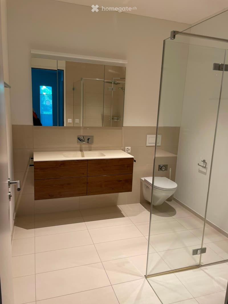 Modern bathroom with white tiled floor, wooden cabinet, sink, toilet, and glass-enclosed shower area.