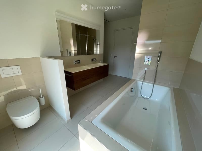 Modern bathroom with a white bathtub, toilet, sink, and large mirror. Features tiled walls and floor, and a sleek wooden vanity with double sinks.