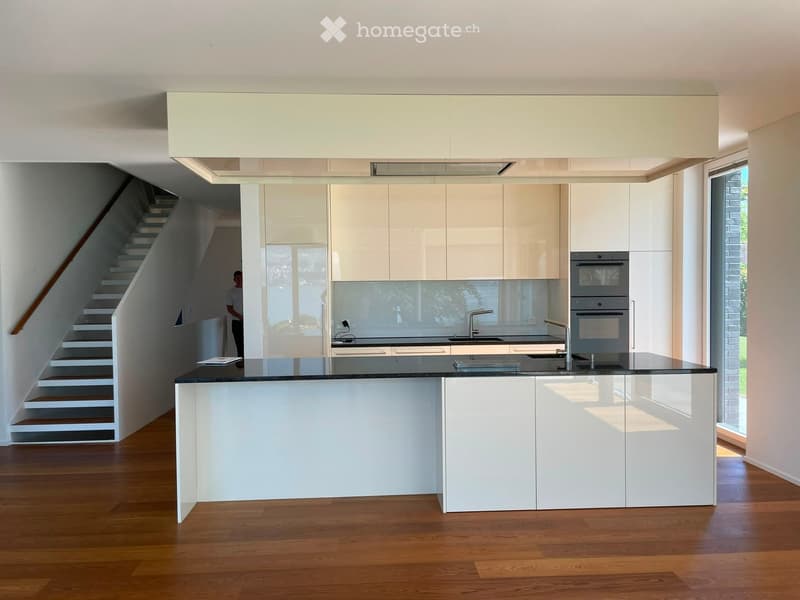 Modern kitchen with white cabinets, a central island, and stainless steel appliances. The space features wooden flooring and a staircase leading to an upper level.