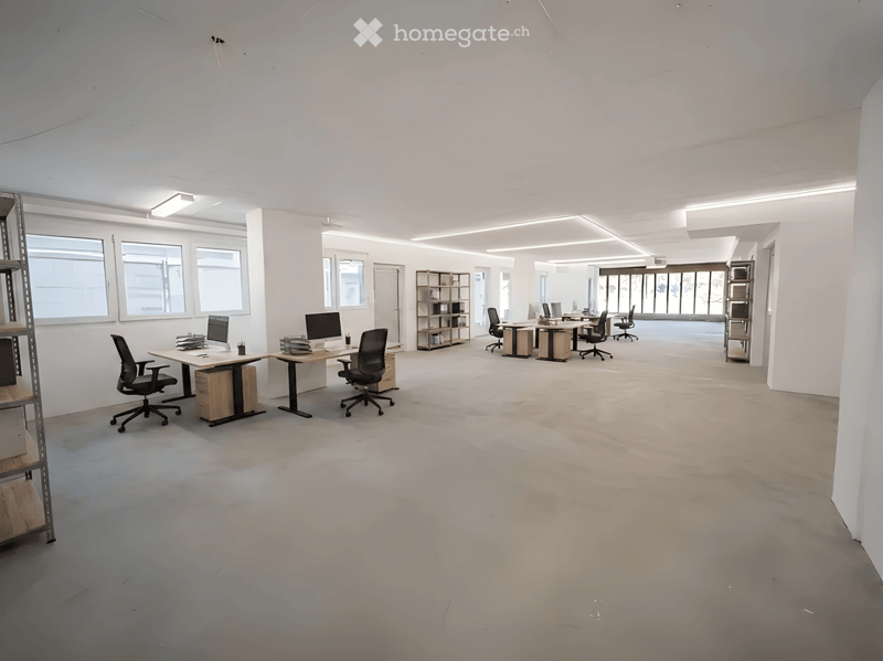 Modern office space with desks, chairs, computers, and shelves, large windows, and neutral color scheme.