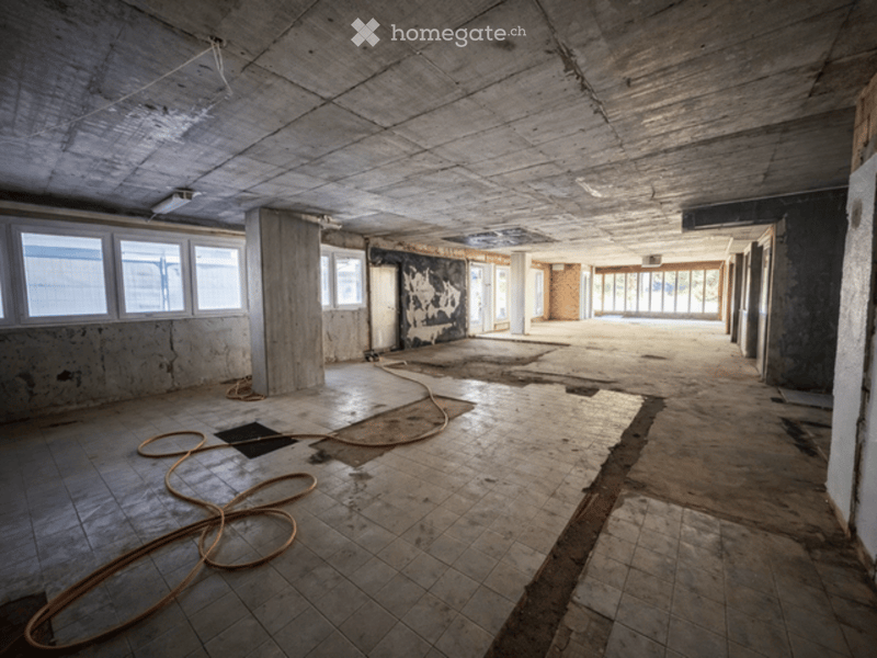 Empty room with exposed concrete ceiling, tiled floor, large windows, and visible structural elements. No furniture or furnishings present.