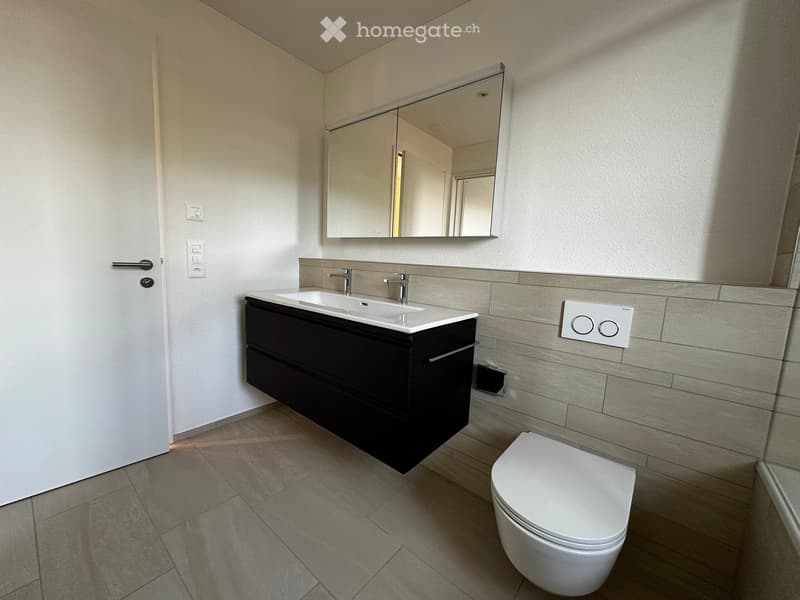 Modern bathroom with a double sink vanity, large mirror, and toilet. The vanity has black lower cabinets and a white countertop. The walls are tiled in a light gray pattern, and the floor is tiled in a similar gray tone.