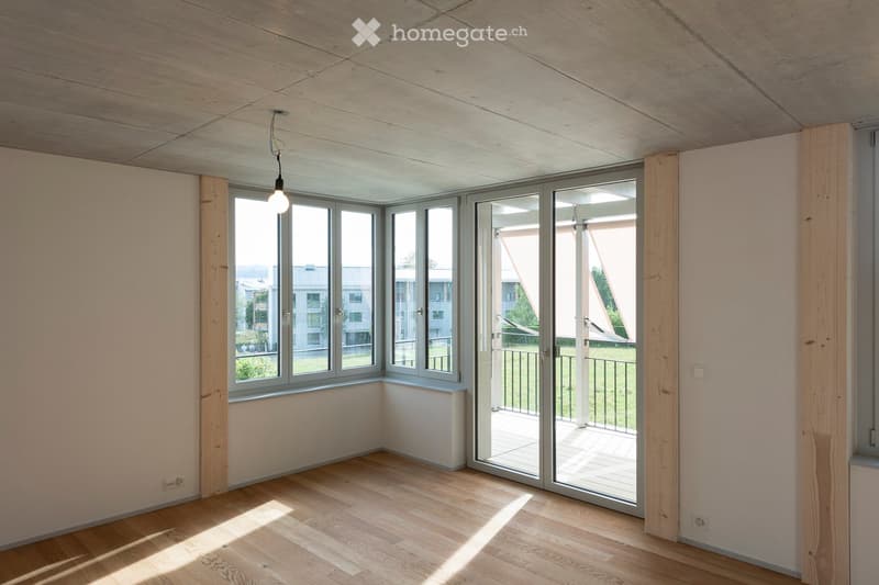 Empty room with wooden flooring, white walls, large windows, and a sliding glass door leading to a balcony.