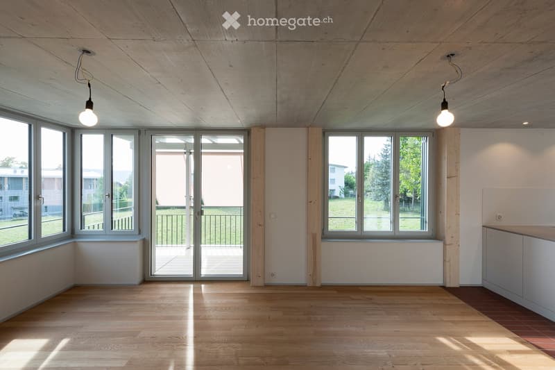 Bright, empty room with wooden flooring, large windows, and a sliding glass door leading to a balcony. Minimalist design with exposed wooden beams on the ceiling and pendant lights.