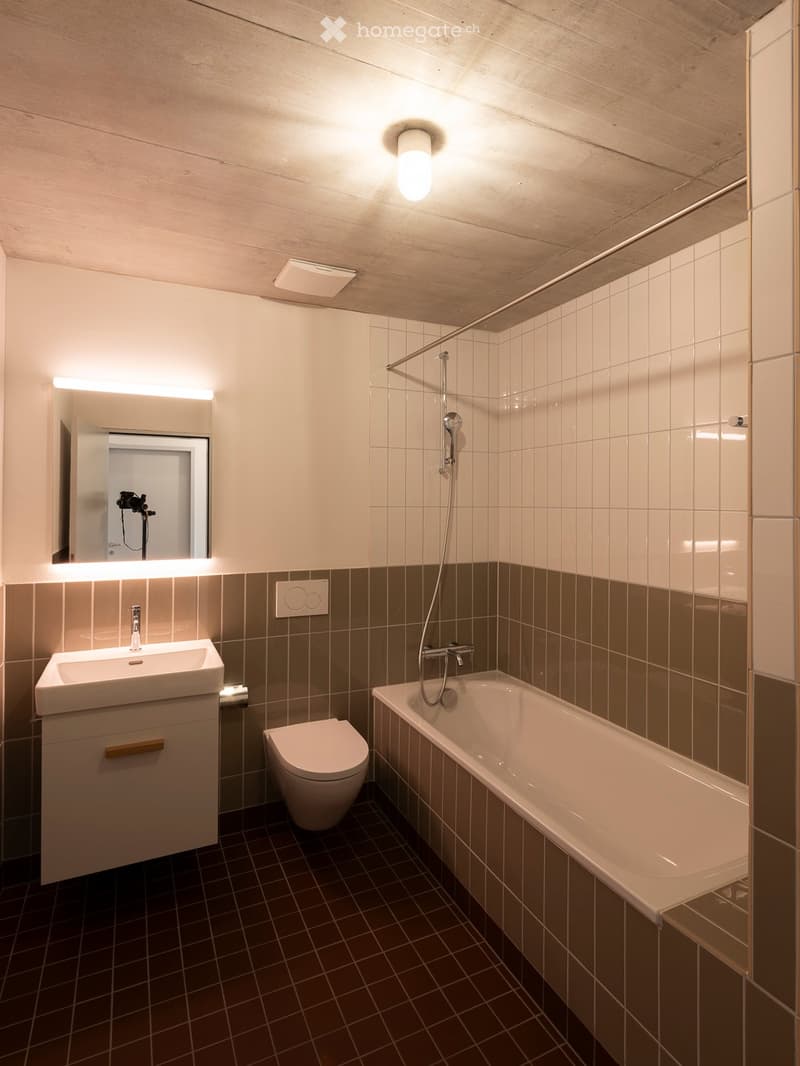 Modern bathroom with a sink, toilet, bathtub, and mirror. Features wooden ceiling, tiled walls, and dark tiled floor.