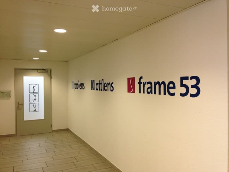 A clean, modern hallway with light-colored walls and a tiled floor. A door with a frosted glass panel is visible on the left, and several company logos are displayed on the right wall.