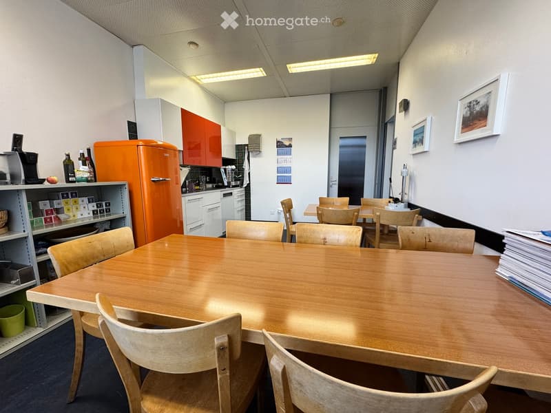 A modern kitchen area with a large wooden table surrounded by chairs, a refrigerator, shelves, and framed pictures on the wall.