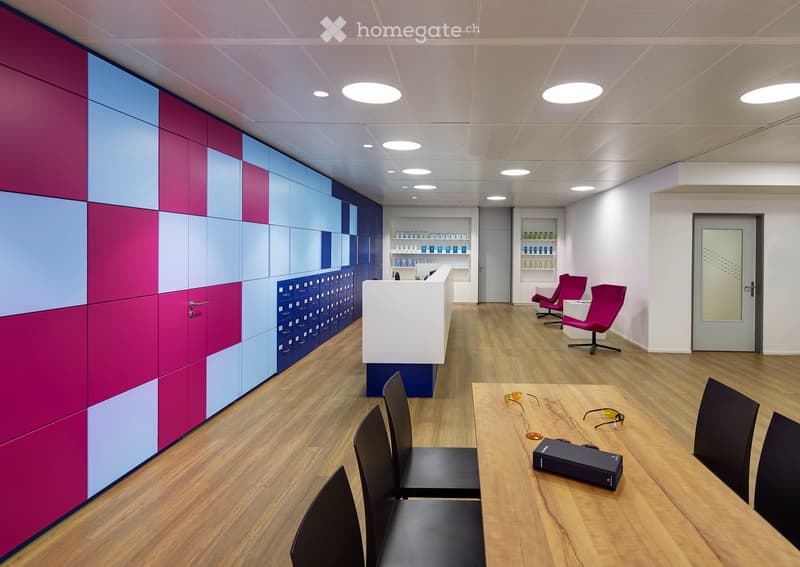 Modern office space with wooden flooring, colorful wall panels, reception desk, chairs, and shelves with items.