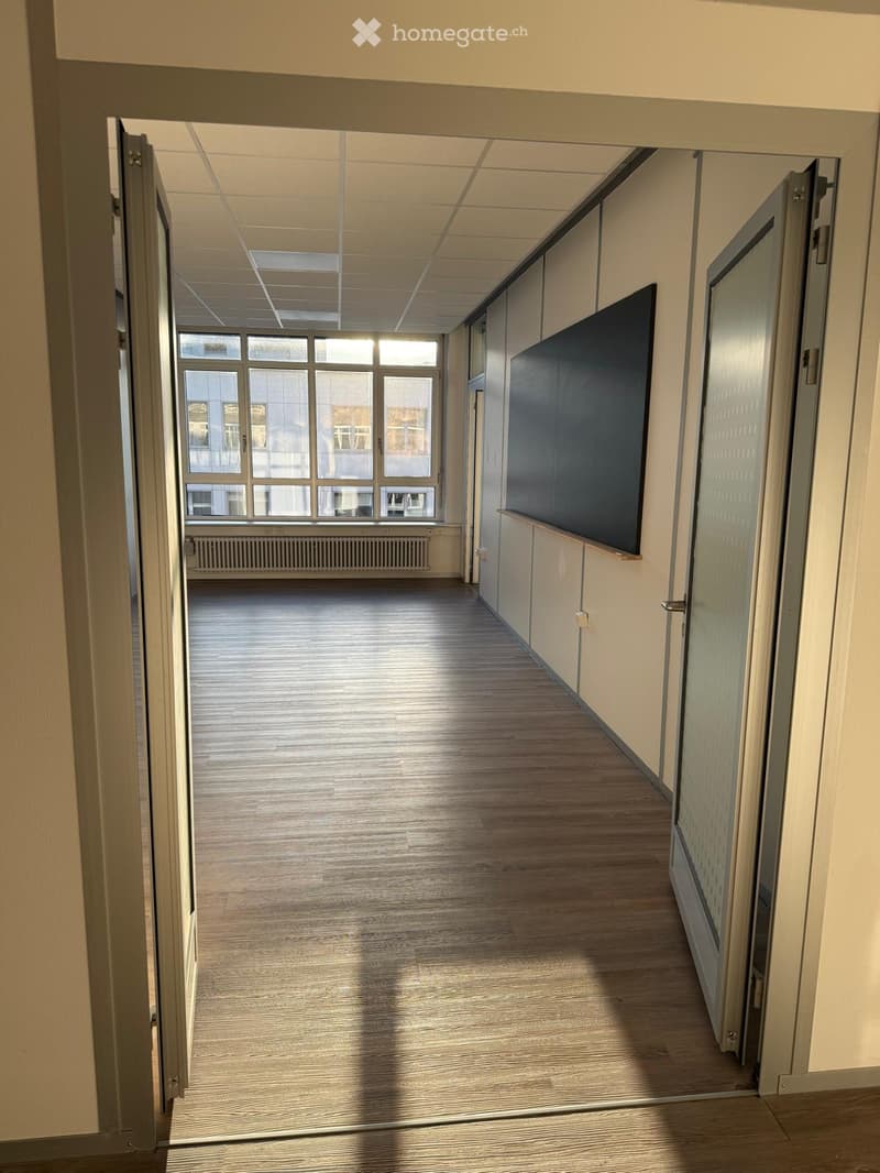 Empty hallway with wooden flooring, large windows on one side, and a radiator below the windows. The walls are painted in neutral tones with a dark panel on one side.