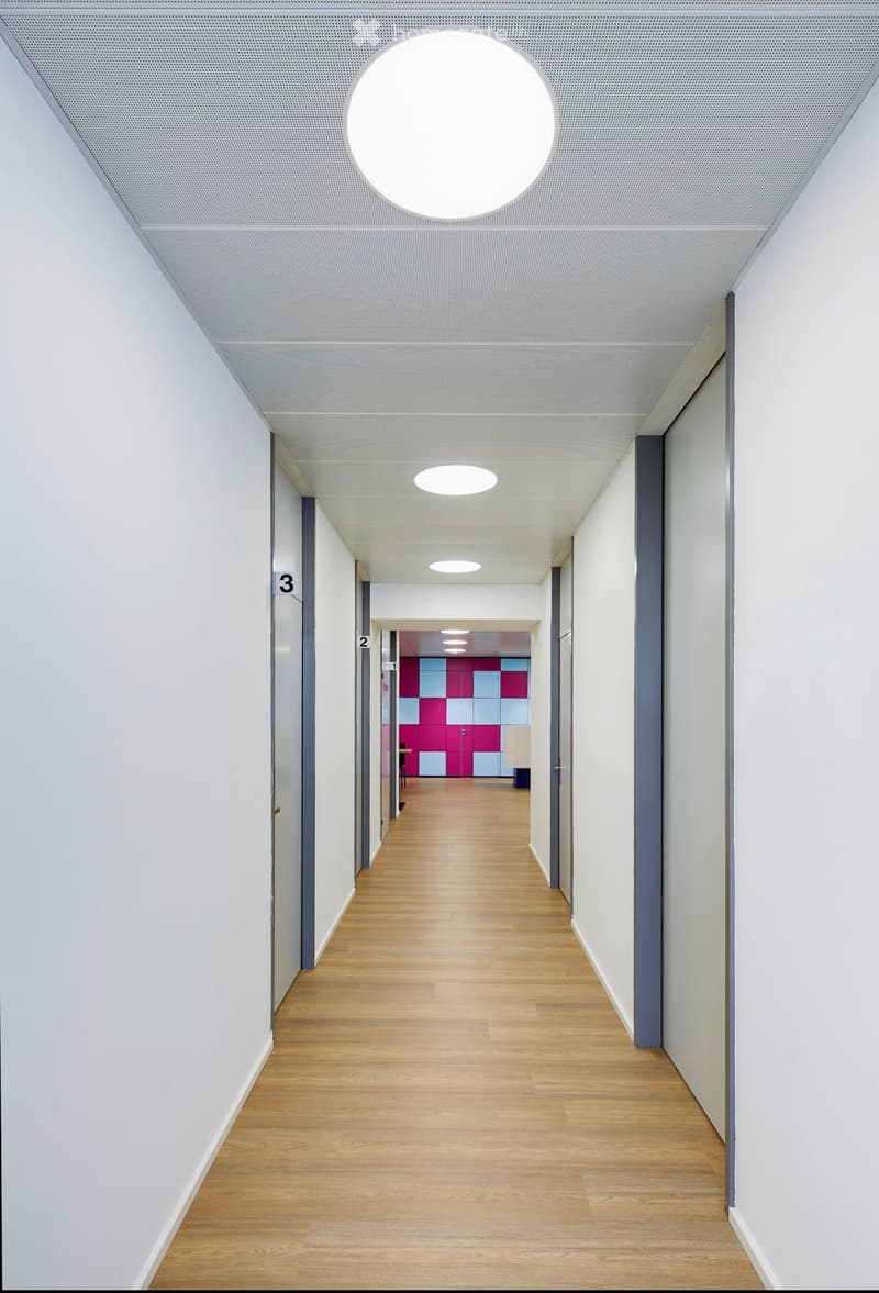 Modern hallway with light wooden flooring, white walls, and recessed lighting. Features numbered doors and a colorful accent wall visible at the end.