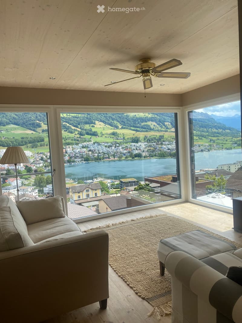 Modern living room with large windows offering a scenic view of a lake and surrounding landscape, featuring a sofa, ottoman, floor lamp, and ceiling fan.