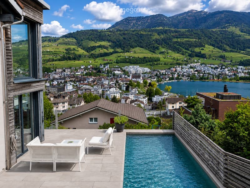 Balcony with a swimming pool, white lounge chairs, a small table, and a scenic view of a town and mountains.