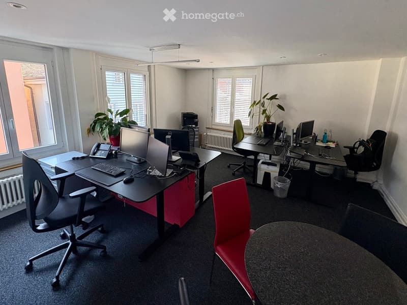 Office space with desks, chairs, computers, printers, and plants. Large windows provide natural light.