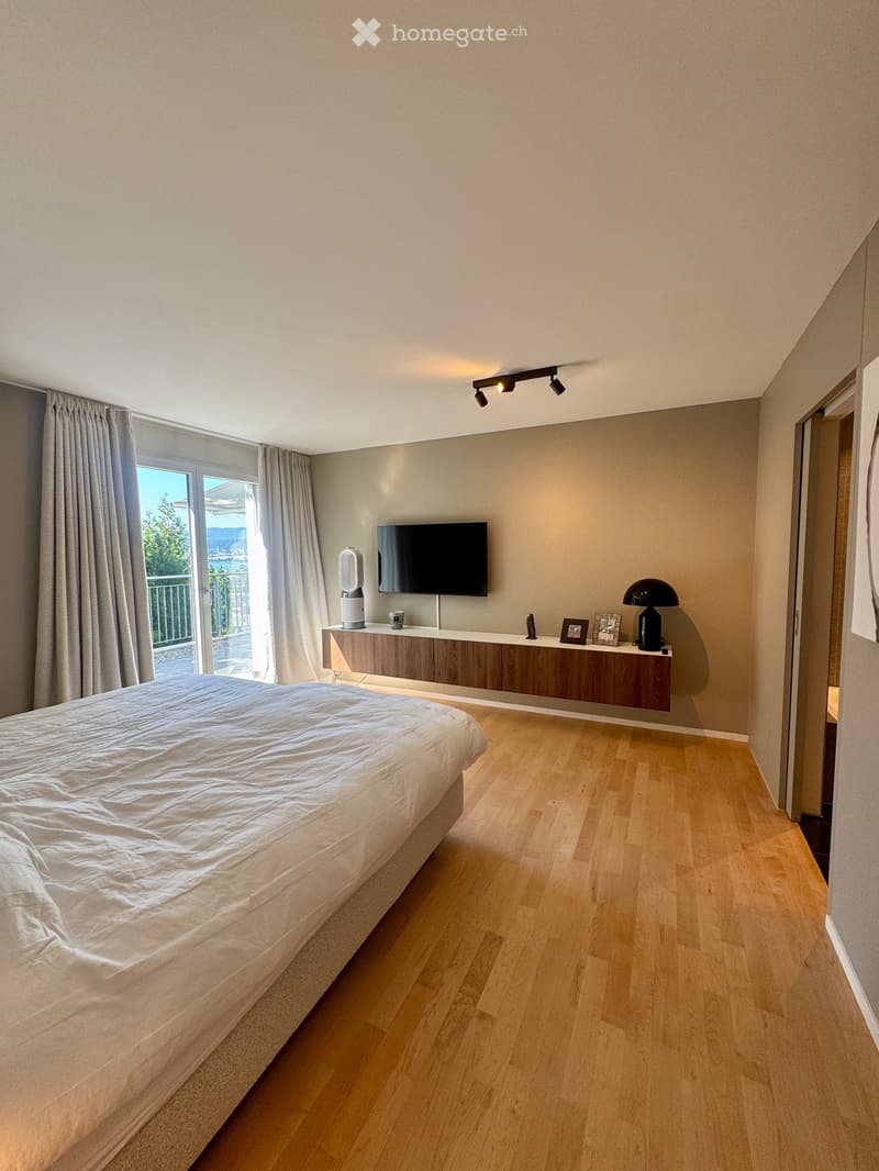 Modern bedroom with a large bed, wooden floor, wall-mounted TV, sleek wooden table, and a black lamp on the table.