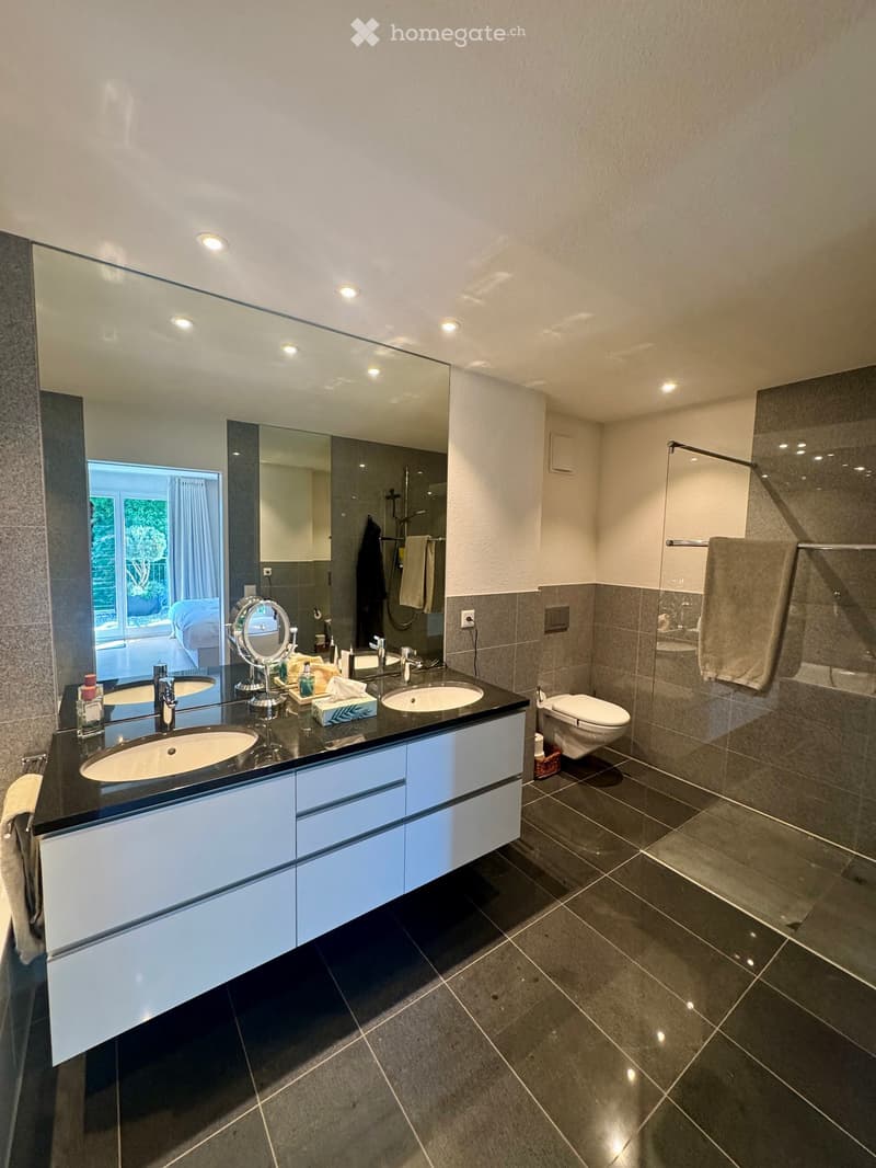 Modern bathroom with double sink vanity, large mirror, toilet, and towel rack. Features dark tiles and recessed lighting.