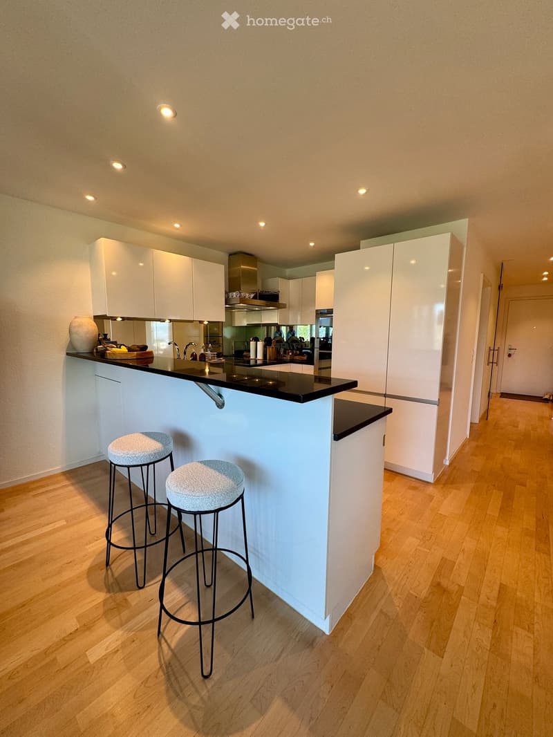 Modern kitchen with white cabinets, black countertop, and two bar stools. Features a central island, built-in appliances, and a sleek design.