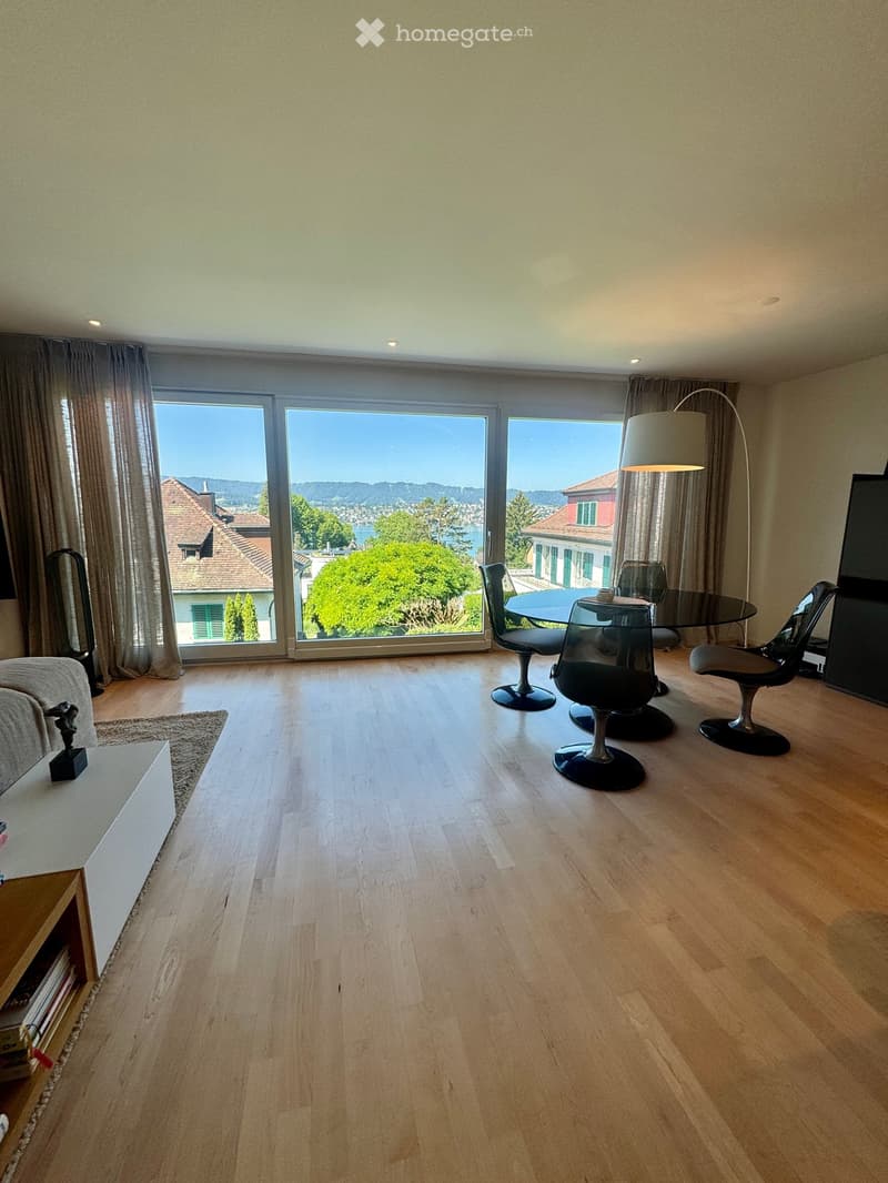 Bright living room with wooden flooring, large windows offering scenic views, sofa, coffee table, dining table with chairs, and a floor lamp.