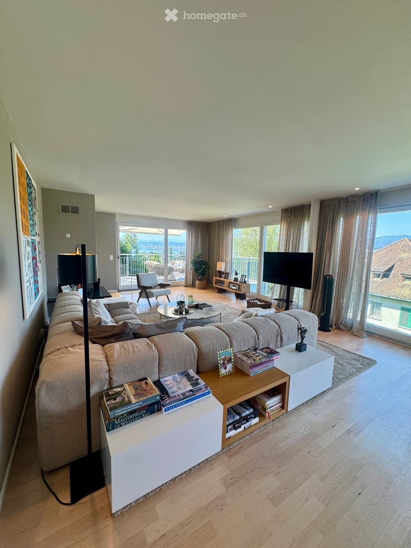 Modern living room with large windows offering a scenic view, featuring a sectional sofa, coffee table, books, and a television.