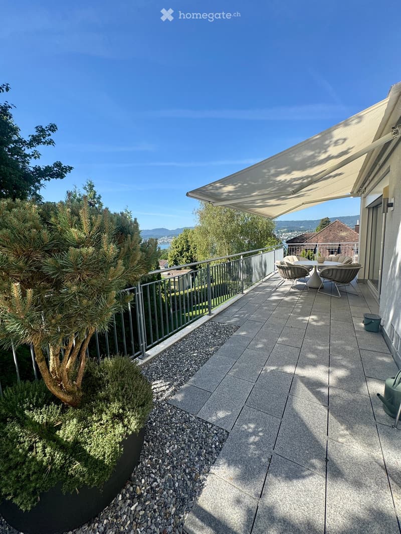 Balcony with tiled floor, metal railing, outdoor furniture including a table and chairs, and a retractable awning.