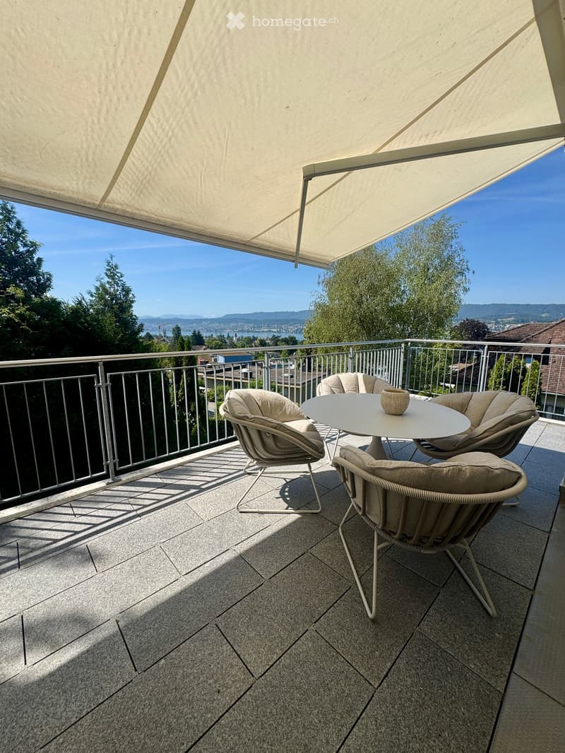 Balcony with a large retractable awning, a round table, four chairs, and a decorative vase, overlooking a scenic view of trees and distant mountains.