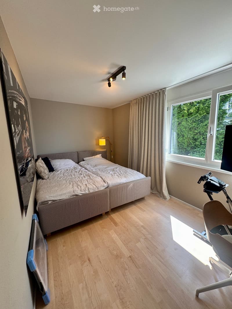 Bright bedroom with wooden flooring, large window with curtains, double bed with pillows and duvet, exercise bike, and a modern lamp on the wall.