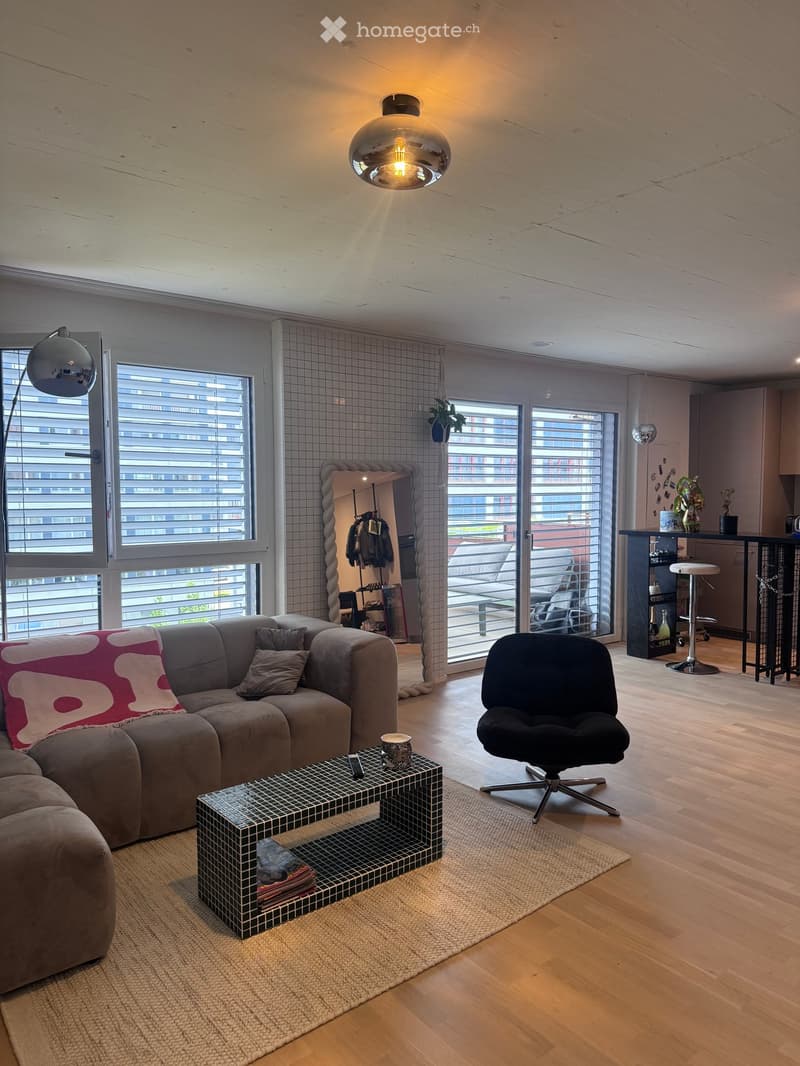 Modern living room with a grey sectional sofa, coffee table, black chair, pendant light, floor lamp, and a view of a balcony area with outdoor furniture.