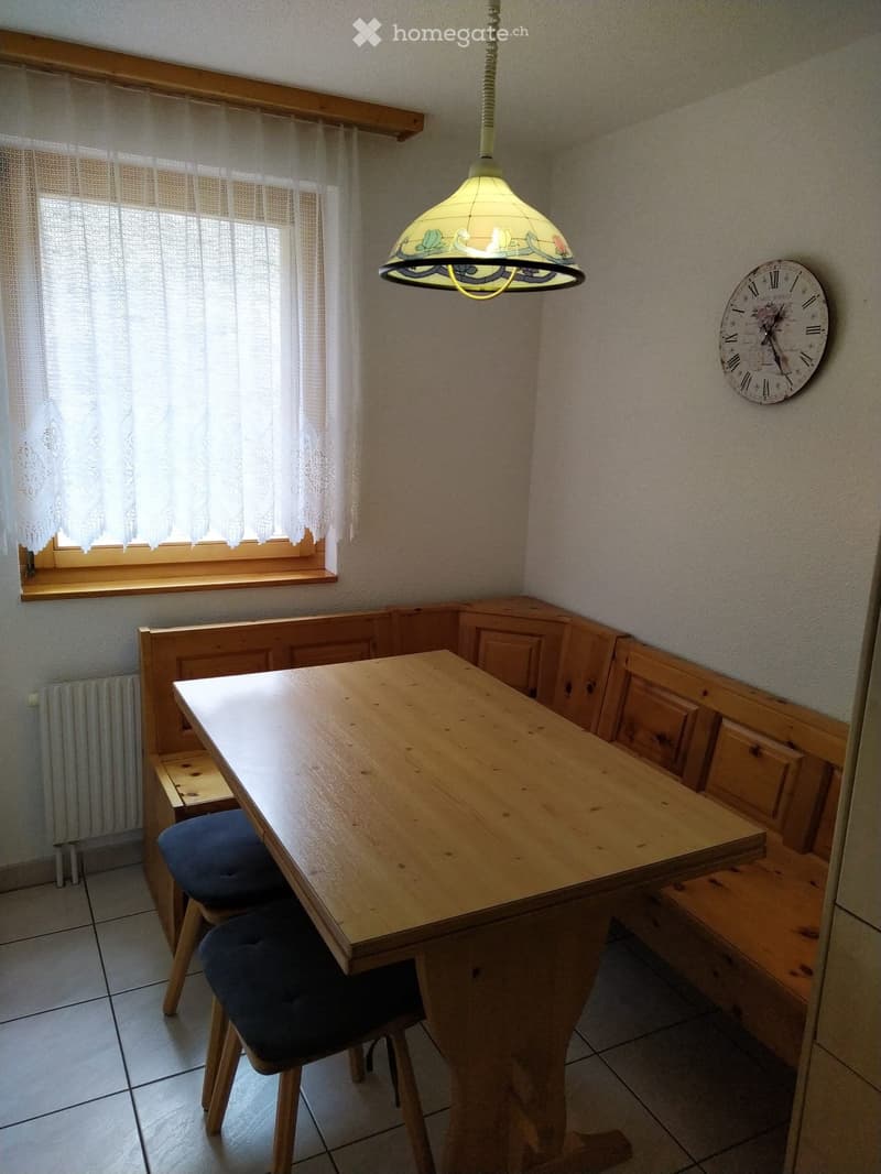 A cozy kitchen with a wooden table, two chairs, a built-in wooden bench seating, a window with white curtains, a decorative hanging lamp, and a wall clock.