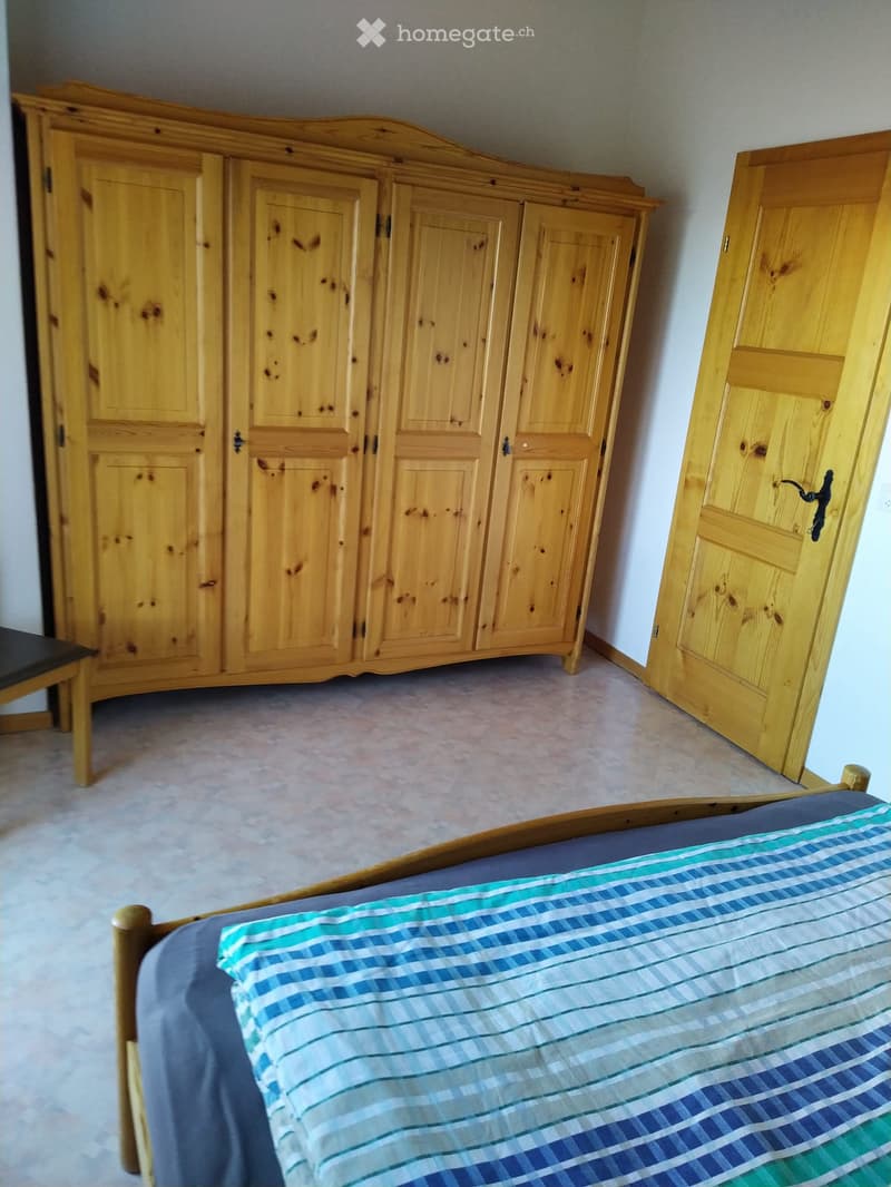 A bedroom with a wooden bed, a large wooden wardrobe, and a small wooden stool. The bed has a blue and green striped duvet cover.
