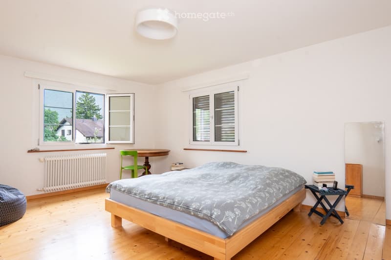 Bright bedroom with wooden flooring, large windows, a bed with a grey duvet, a small table with a green chair, and a stool with books.